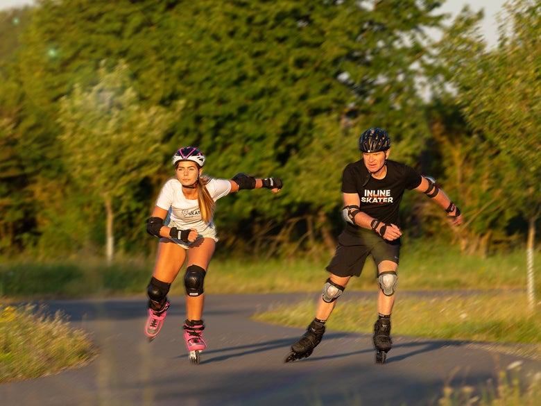 A female and male inline skating on a sunny day with plenty of greenery, wearing full protective gear