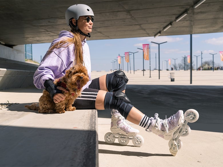 A girl and a dog sitting on a curb. She is wearing the Powerslide Onsie Knee Pads, a helmet and a pair of inline skates.