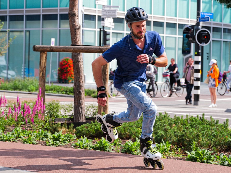 A man rollerblading energetically on a city path, wearing jeans, a blue shirt, and a helmet. Bright, sunny day with cyclists in the background.