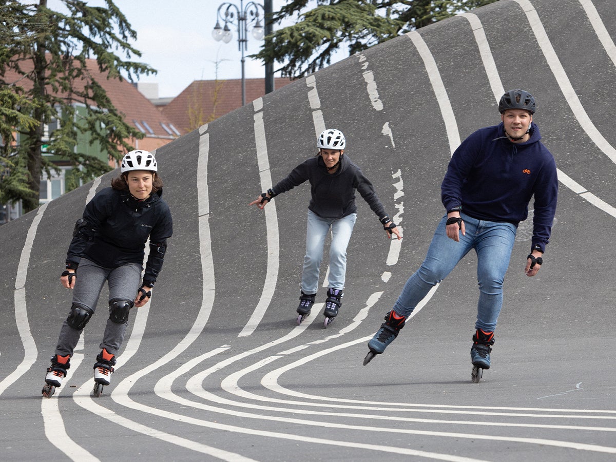 Three people rollerblading downhill on a curved, striped concrete surface, wearing helmets and casual outfits, conveying excitement and fun.