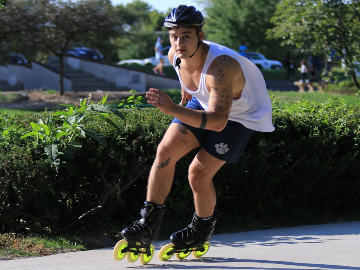 A man on inline fitness skates moves swiftly on a sunny park path, wearing a helmet, white tank top, and blue shorts. Tattoos are visible on his arms and legs, conveying an athletic and energetic vibe.
